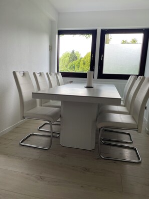 A modern dining area with a white table and chairs, large windows with a view of greenery, and a radiator under the table. - Blauer Stein - Pure - bl07-haus (Pulheim)