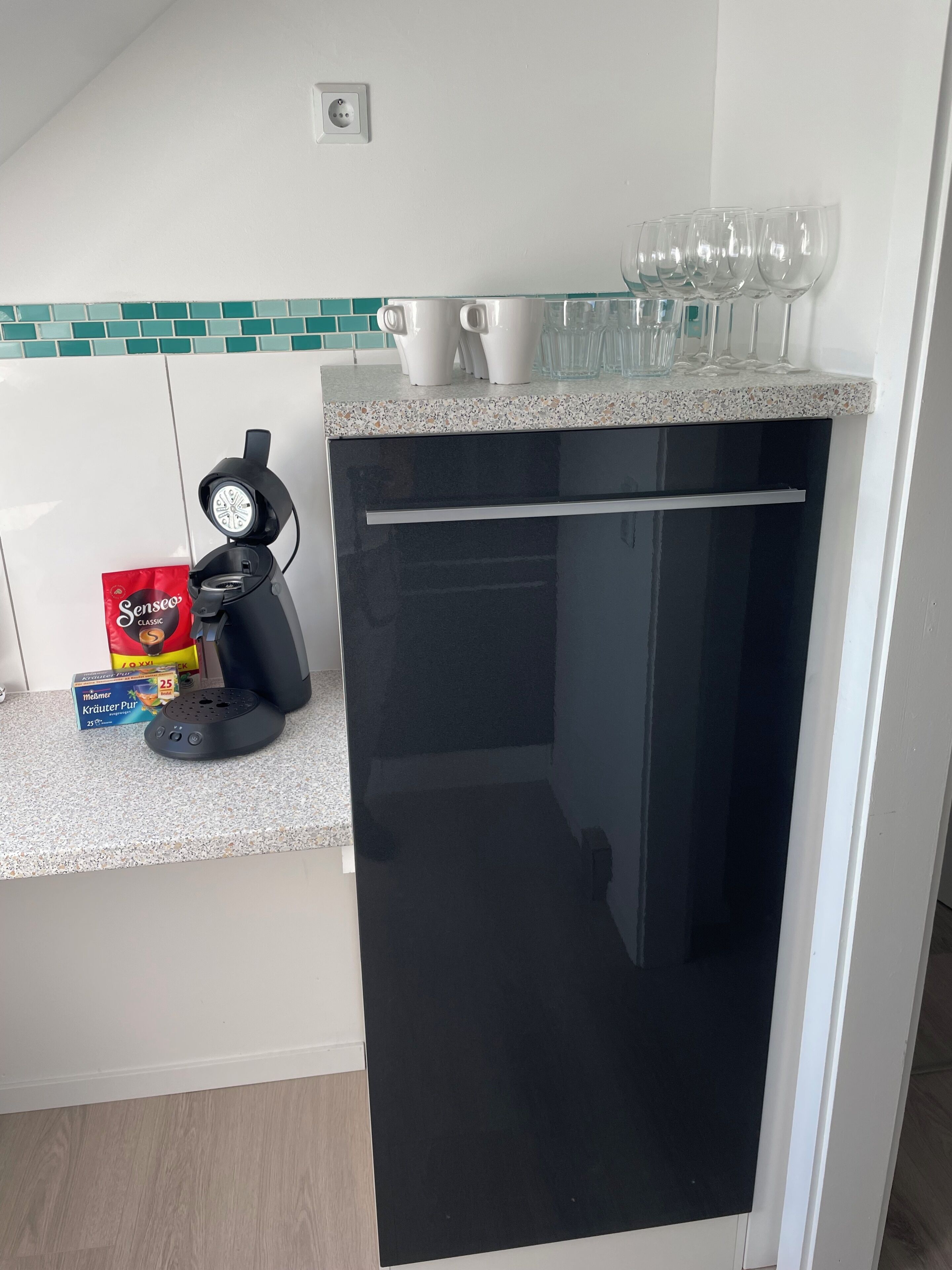 A kitchen with a coffee machine, a box of coffee pods, and a set of wine glasses on the counter.