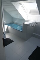 Un tragaluz sobre una bañera en un baño con azulejos blancos y un perchero metálico.