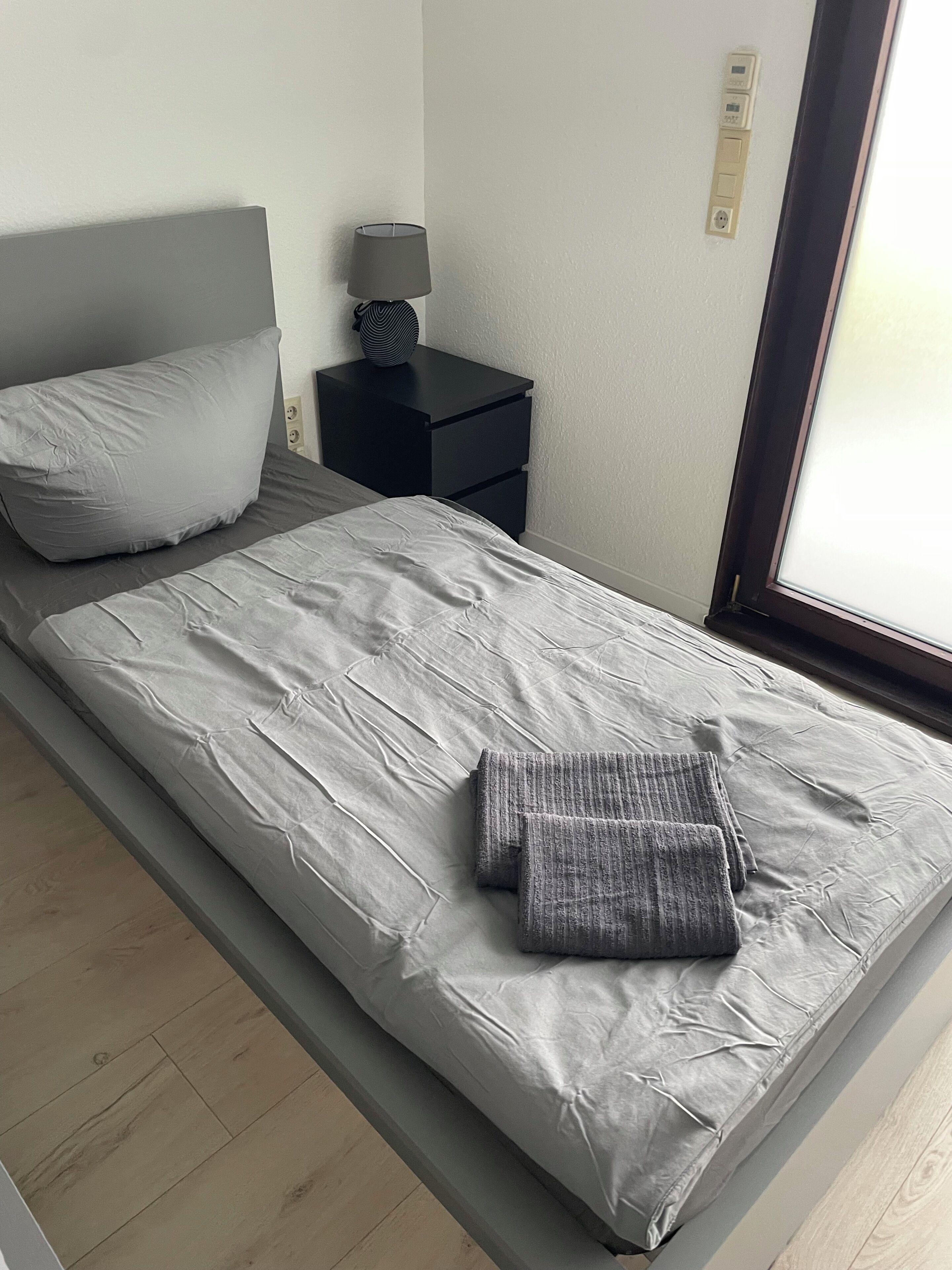 Ein Einzelbett mit grauer Matratze und Kissen, ein kleiner Nachttisch mit Lampe und ein Fenster mit Jalousien.
