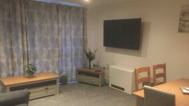 Una habitación con un televisor montado en la pared, una mesa de comedor de madera con sillas, un escritorio pequeño de madera y una planta en maceta.