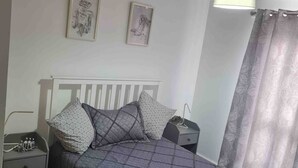 Un dormitorio con cama, mesita de noche, lámpara y cortina.