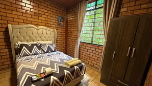 A bedroom with a brick wall, a bed with a patterned bedspread, a wooden wardrobe, and a window with curtains. - Country House in Tingo Maria - Adelaide Resort (Tingo María)