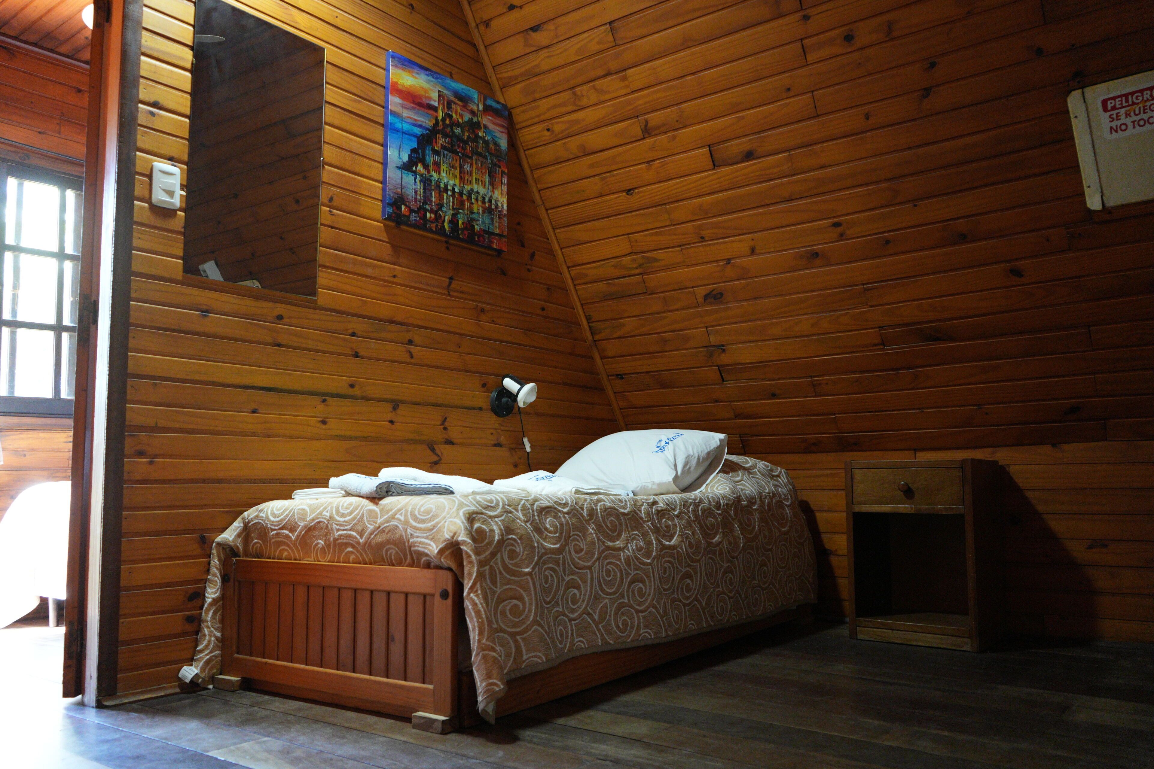 Quarto de cabana de madeira com uma cama, um criado-mudo, uma luminária na parede e um quadro.
