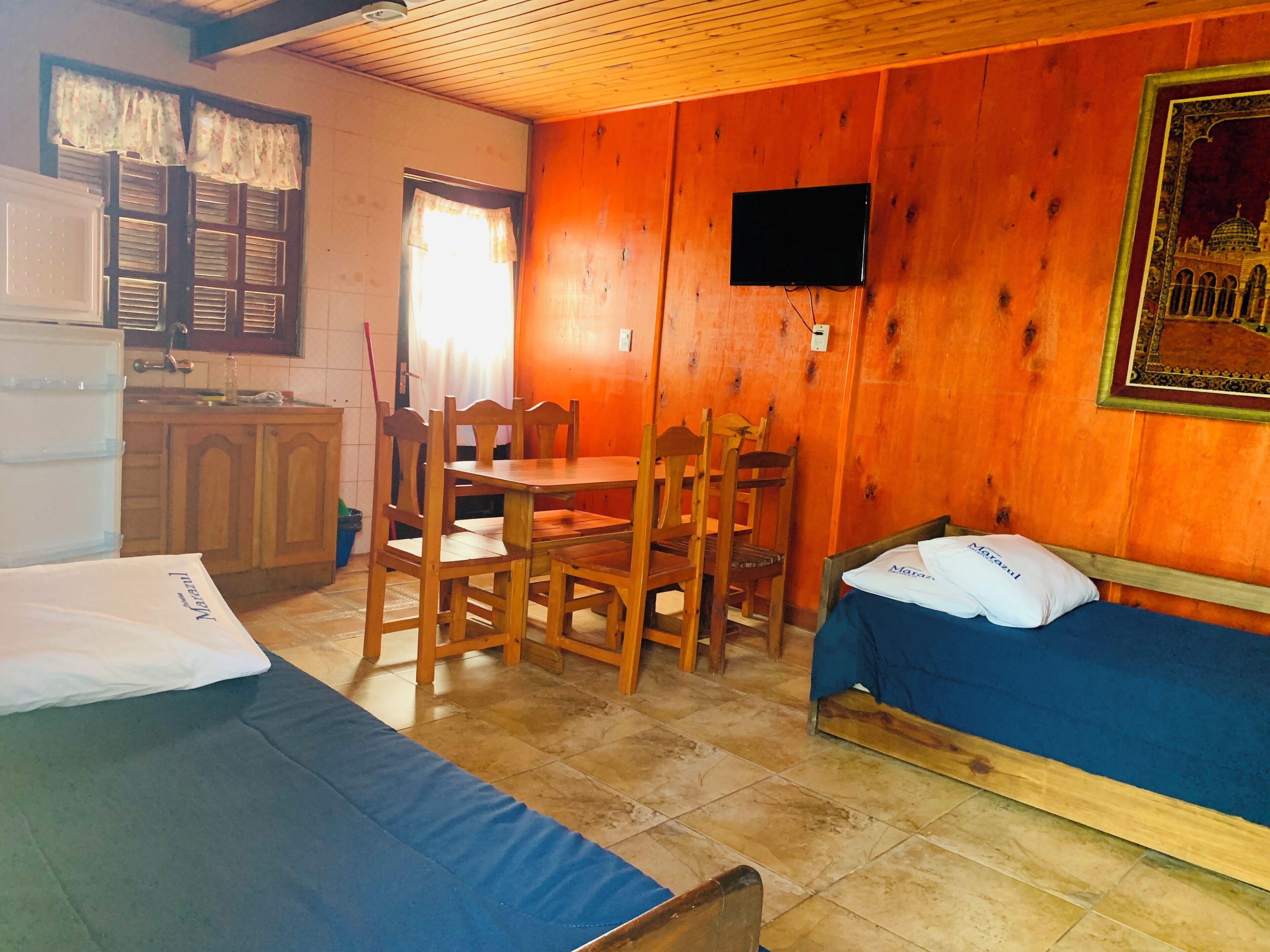 Una habitación con cama, una mesa con sillas, un televisor montado en la pared y un ventanal con cortinas.