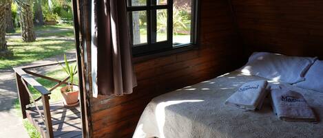 Una habitación de madera con una cama, una ventana con cortinas, una planta en maceta y una terraza de madera.