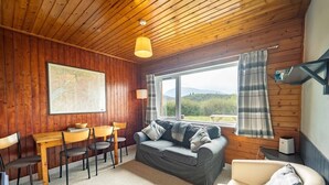 Een gezellige kamer met een houten plafond, een bank, een eettafel met stoelen en een groot raam met uitzicht op de omgeving.