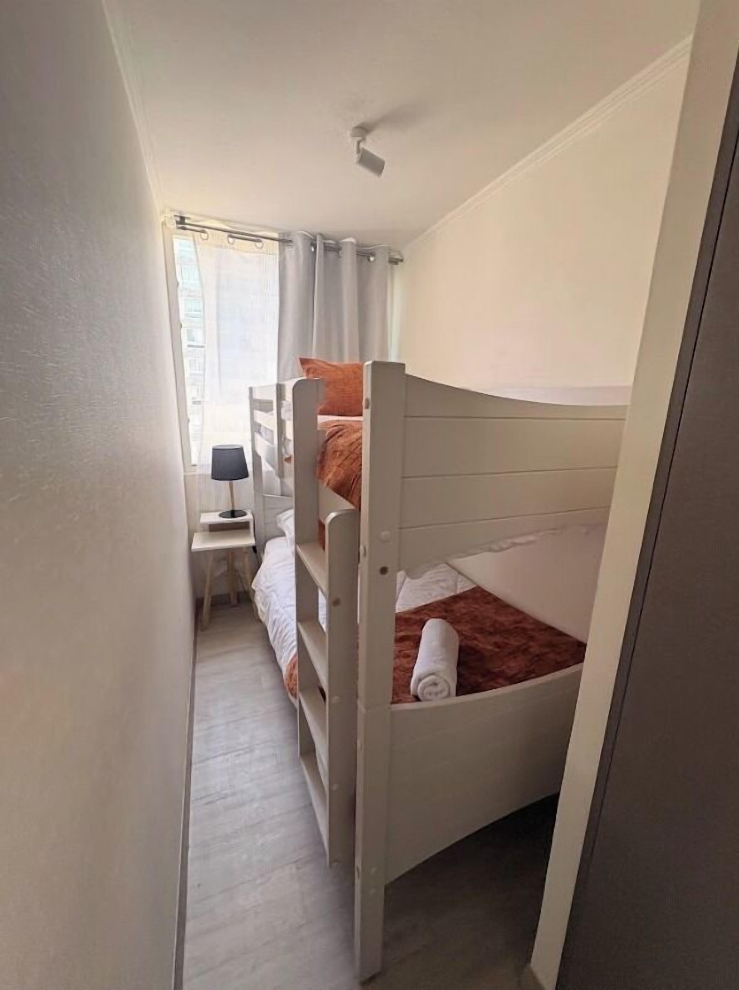 Quarto pequeno com duas camas beliche, uma mesinha de cabeceira com abajur e uma janela com cortinas.