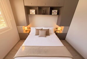 A neatly made bed with two pillows, a bedside table with a lamp, and a shelf above the bed with framed pictures.