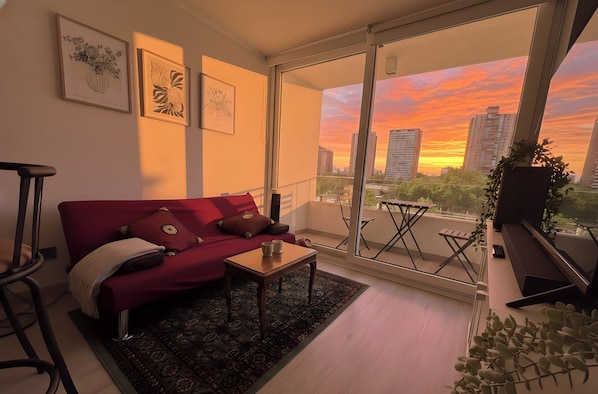 A modern living room with a red sofa, a small wooden table, and a view of a sunset over a cityscape.