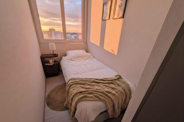 A small, neatly arranged bedroom with a single bed, a bedside table with a lamp, and a view of the city through a window. - Edificio Accion Maule by Domus Travels (Santiago)