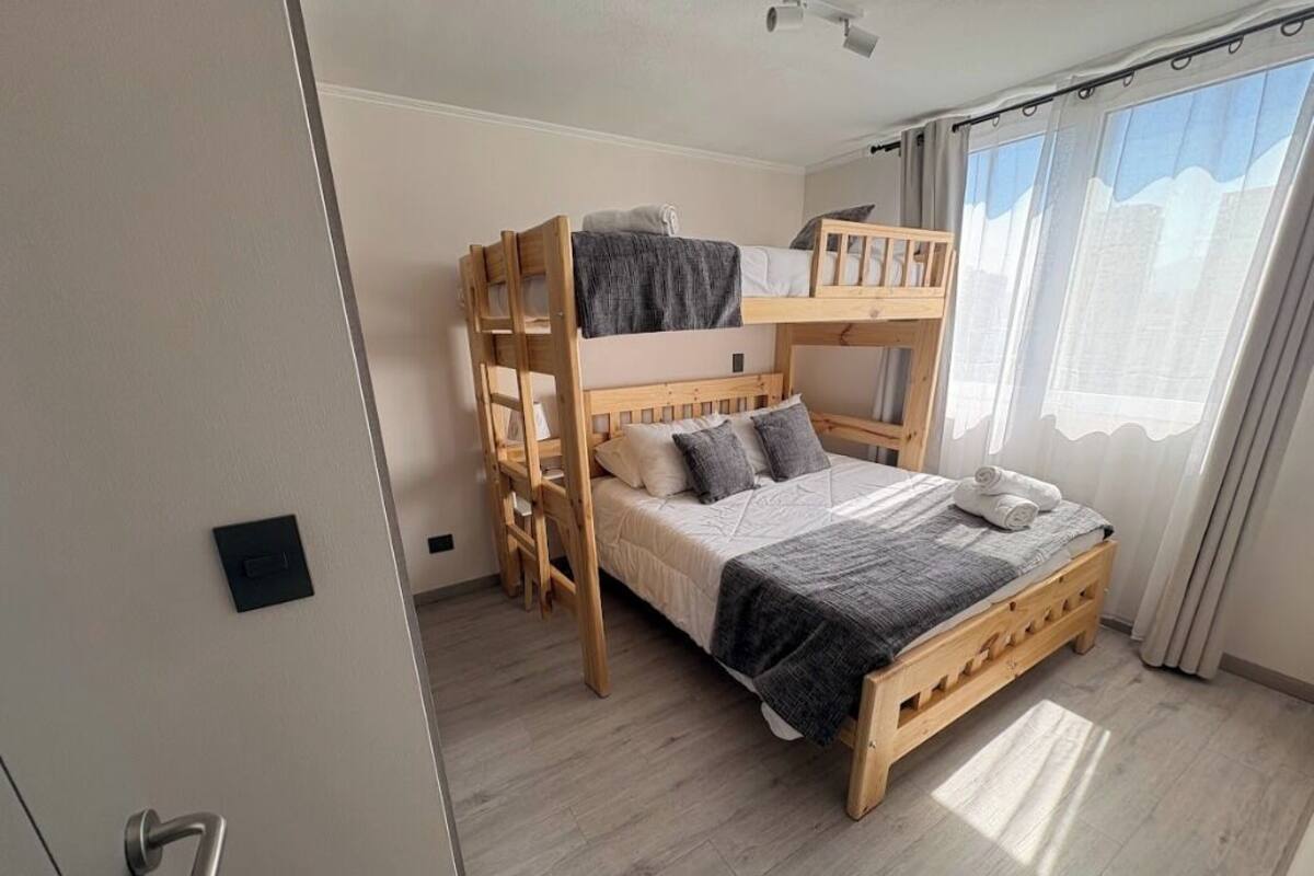 Habitació amb un llit superposat, una escala de fusta, una taula de nit i una finestra amb cortines.