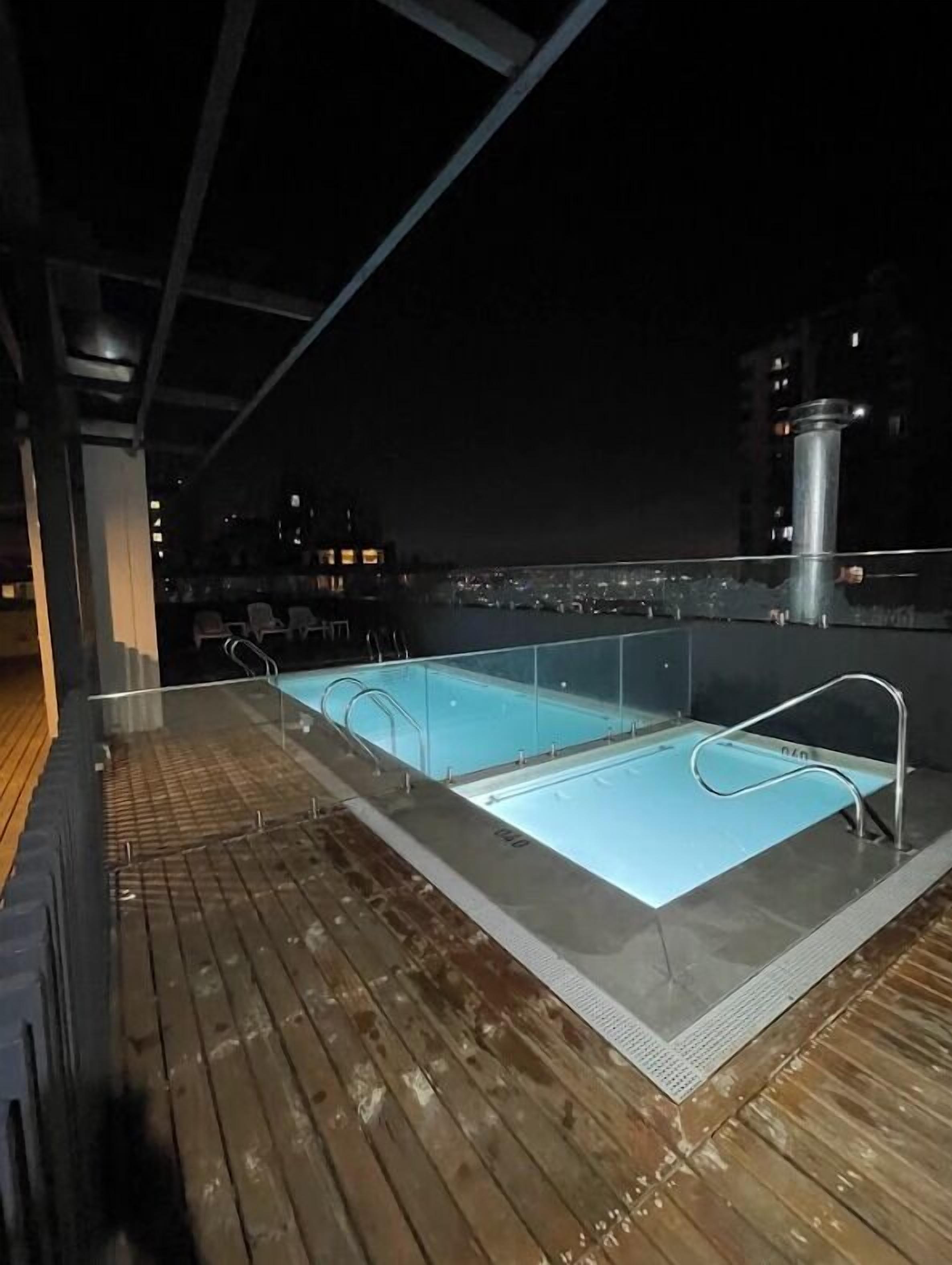 A rooftop pool area with clear glass railings, a wooden deck, and a city view at night.