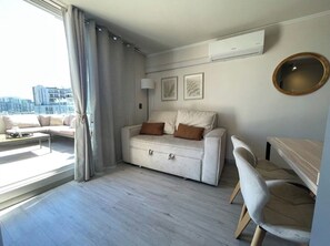A modern living room with a sofa, a coffee table, and a desk. There is a large window with curtains, a wall-mounted air conditioner, and framed artwork.
