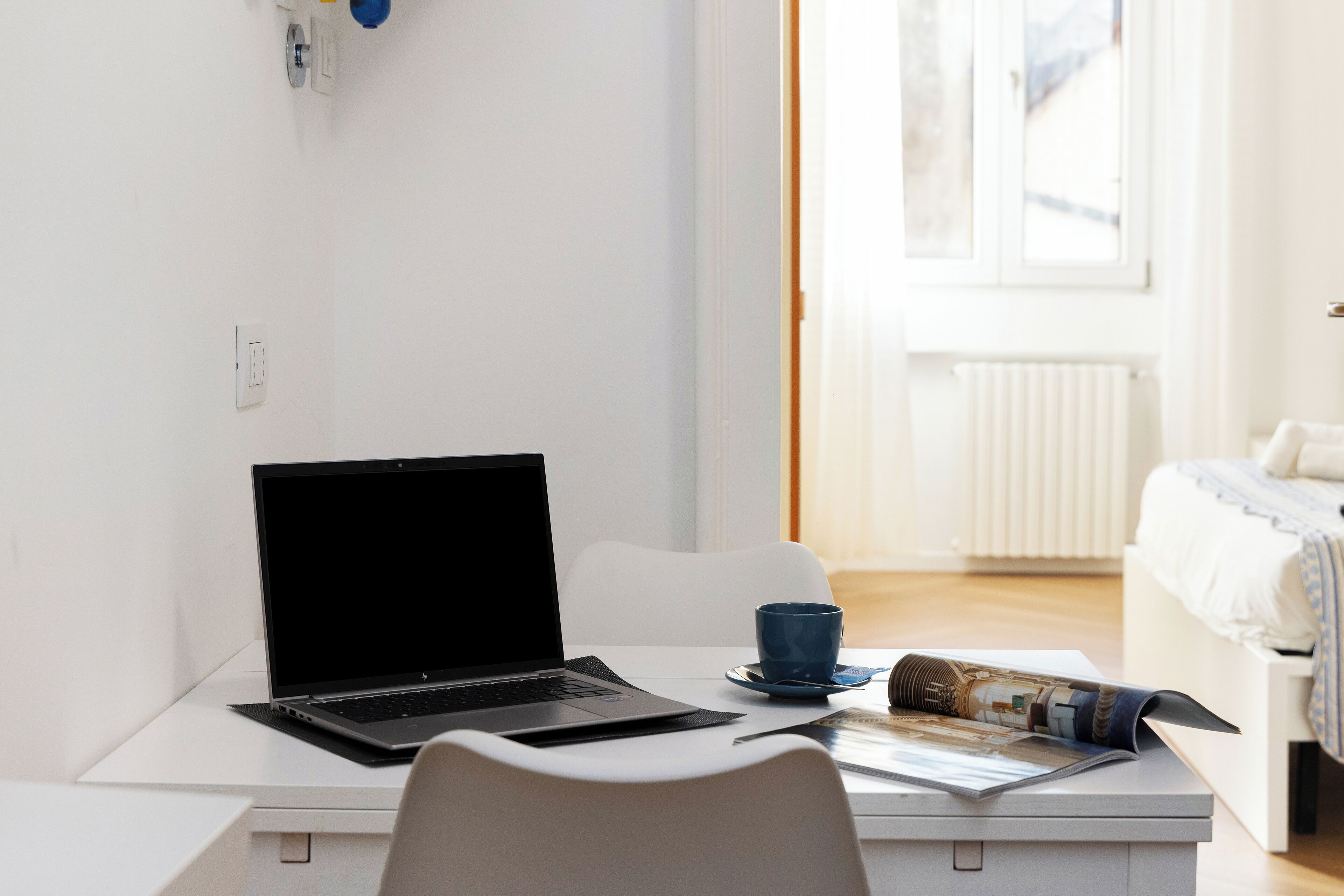 A laptop on a white desk with a blue mug and a rolled-up magazine.