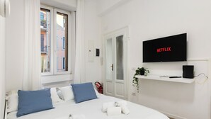 Ein modernes Hotelzimmer mit einem Bett, einem Fernseher, der 'NETFLIX' anzeigt, einer kleinen Ablage mit einer Pflanze und einem Fenster mit Vorhängen.