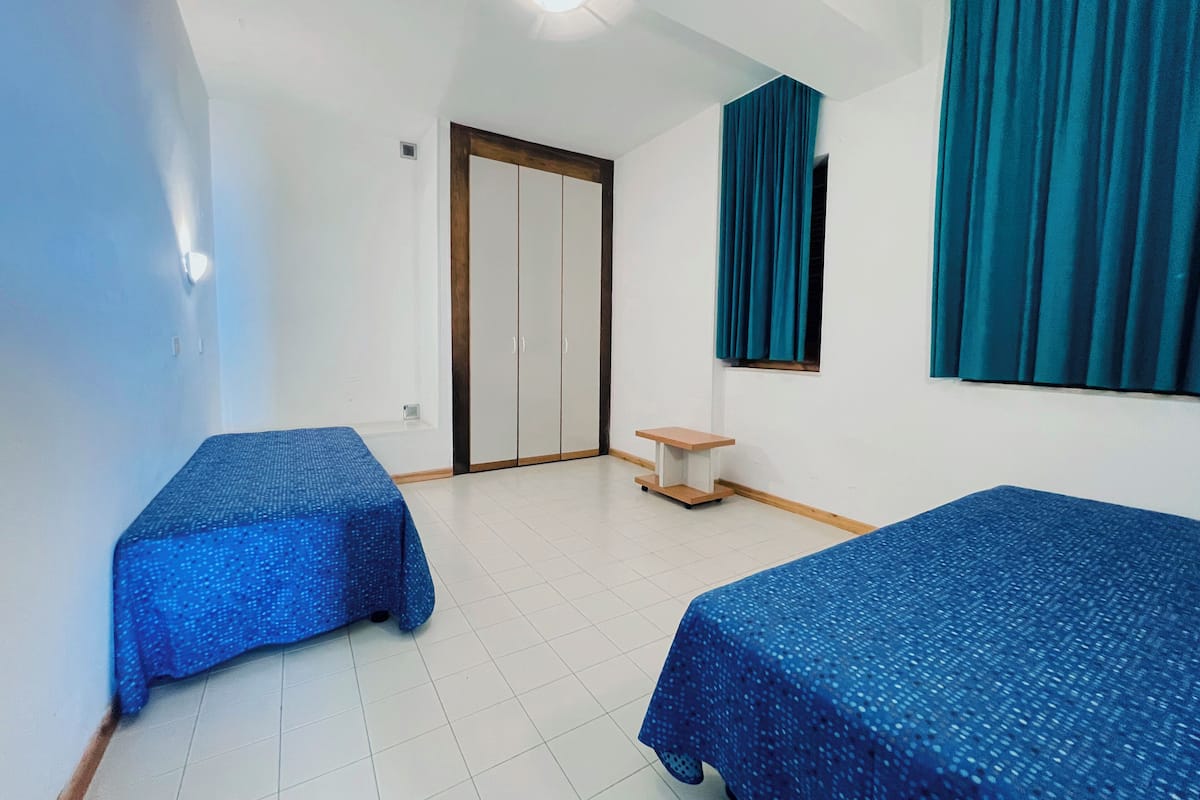 A room with two single beds, a small table, a wardrobe, and a window with curtains.