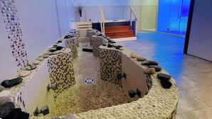 Uma área de spa com piso de mosaico de pedras, um espaço de estar central com um elemento aquático e uma escadaria que leva a um ambiente envidraçado.