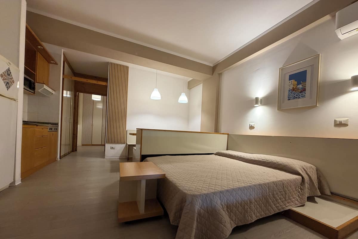 A bedroom with a bed, bedside table, and a view of the kitchen and bathroom.