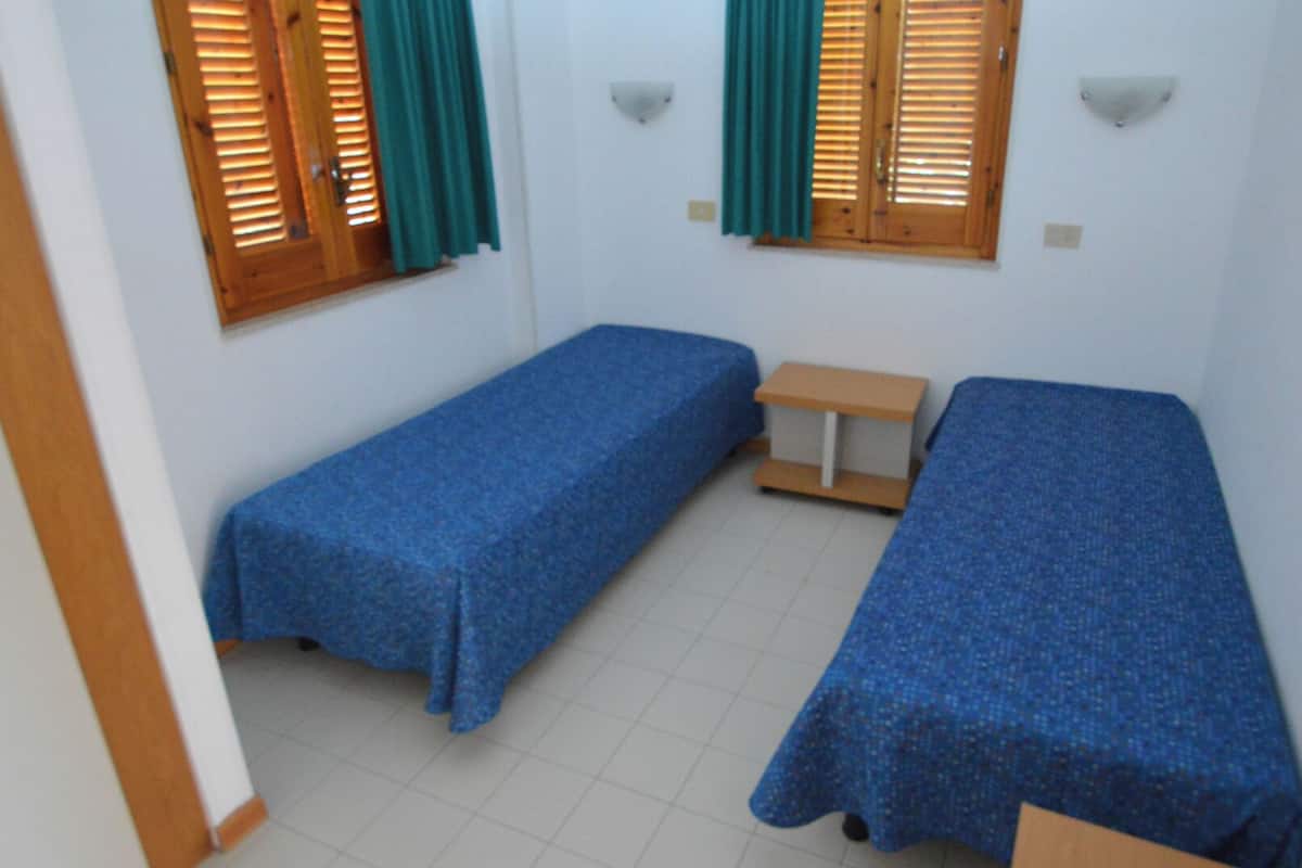 A small room with two single beds, a small table, and two windows with wooden shutters and green curtains.
