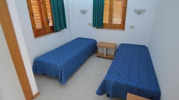 A small room with two single beds, a small table, and two windows with wooden shutters and green curtains.