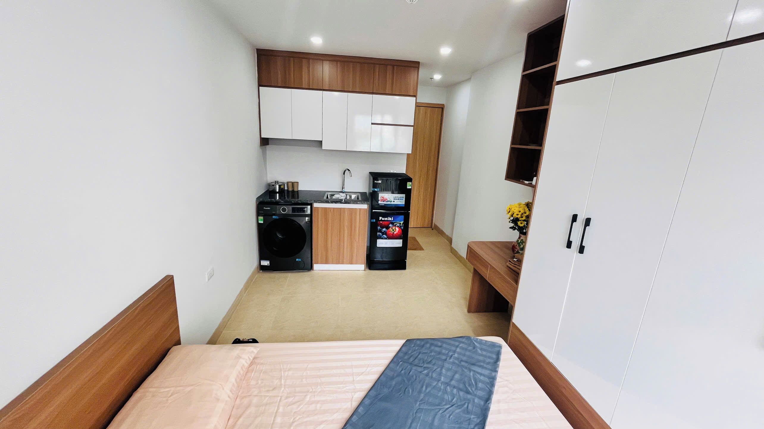 Un dormitorio pequeño y moderno con una cama, una mesita de noche de madera y un armario. En el fondo, se aprecia una kitchenette compacta con lavadora, un refrigerador pequeño y un lavamanos.