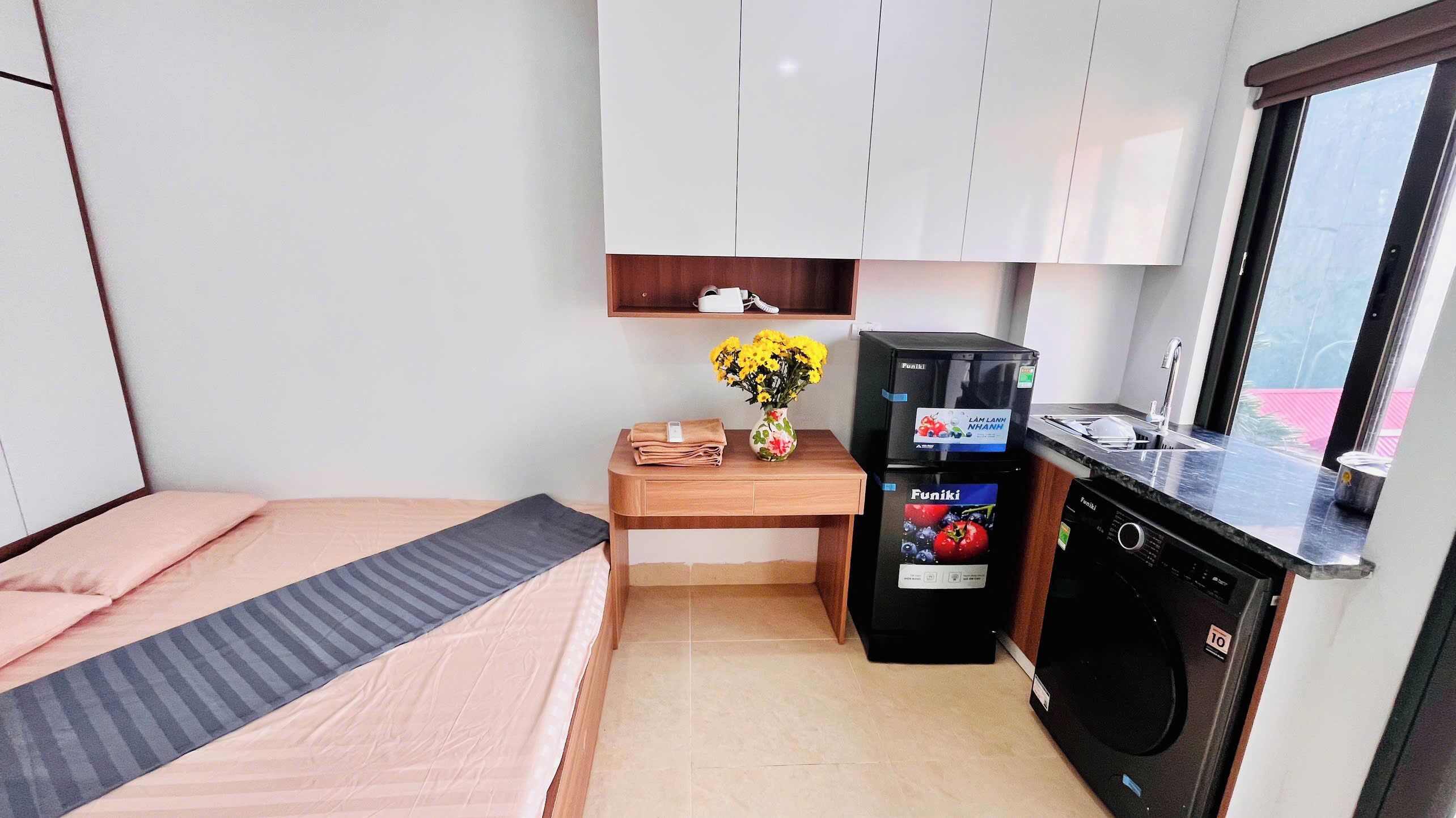 Une chambre d’hôtel compacte comprenant un lit, une petite table en bois ornée d’un vase de fleurs, un minibar et une kitchenette équipée d’un évier et d’une cuisinière.