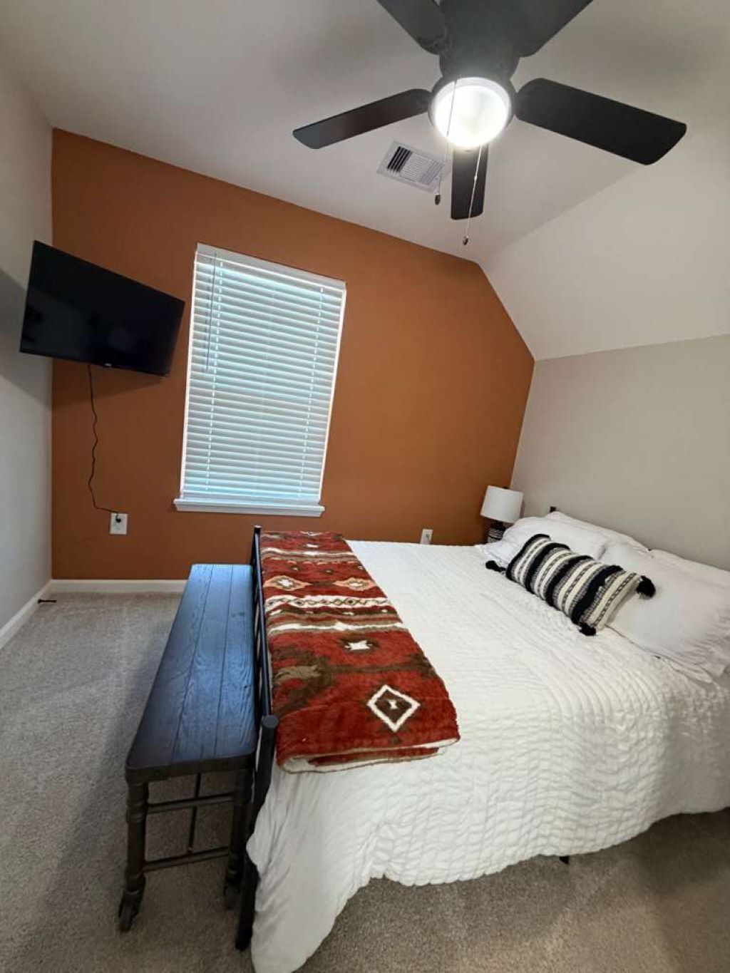 Ein Schlafzimmer mit Bett, Schreibtisch, Deckenventilator, Wand-Fernseher und einem Fenster mit Jalousien.