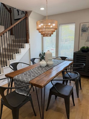 Un comedor con una mesa de madera, sillas negras, una lámpara colgante tejida y una escalera con un letrero en forma de 'Y'.