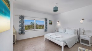 Une chambre à coucher avec un lit, des tables de chevet, une fenêtre avec des rideaux, un tableau et une vue sur le paysage.