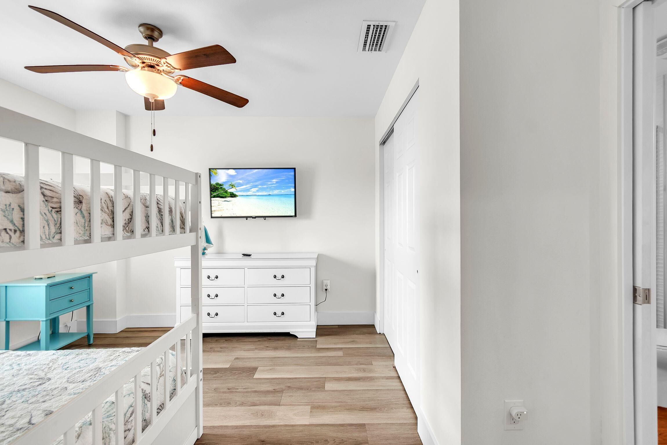 A bedroom with a wooden floor, white walls, a ceiling fan, a TV on the wall, a white dresser, and a blue nightstand.