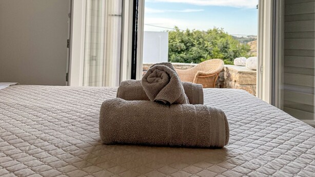 Una cama con cubierta acolchada y dos toallas enrolladas, una puerta abierta que da a un balcón con vistas a la vegetación y un cielo despejado.