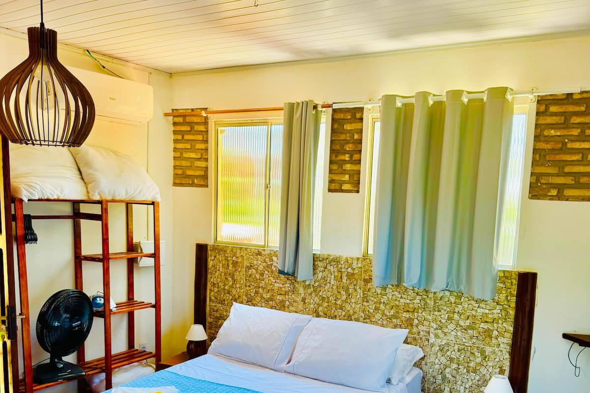 A hotel room with a bed, a bedside table, a fan, and a window with curtains.
