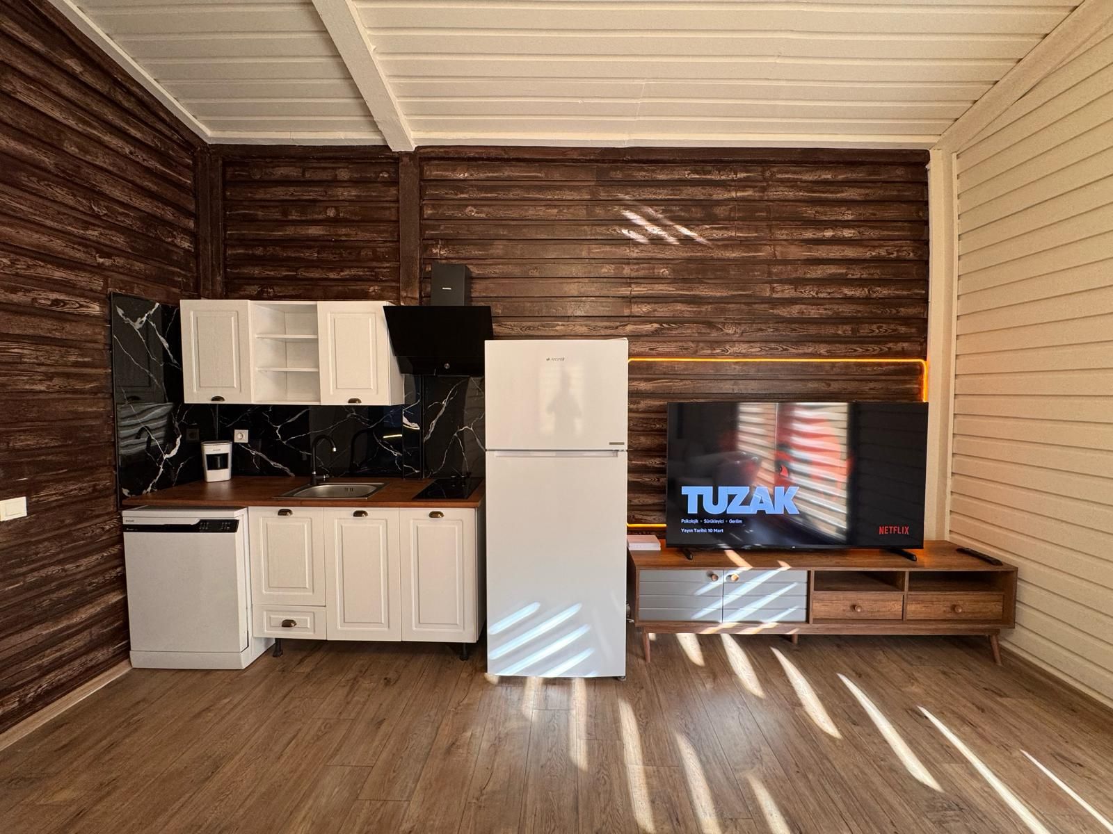 A compact kitchen with white cabinets, a black marble backsplash, a white refrigerator, and a wooden cabinet with a television displaying 'TUZAK' on the screen.
