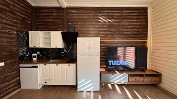 A compact kitchen with white cabinets, a black marble backsplash, a white refrigerator, and a wooden cabinet with a television displaying 'TUZAK' on the screen.