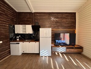 Une cuisine compacte avec des meubles blancs, un pare-feu en marbre noir, un réfrigérateur blanc et un meuble en bois sur lequel est posée une télévision dont l’écran affiche le mot « TUZAK ».