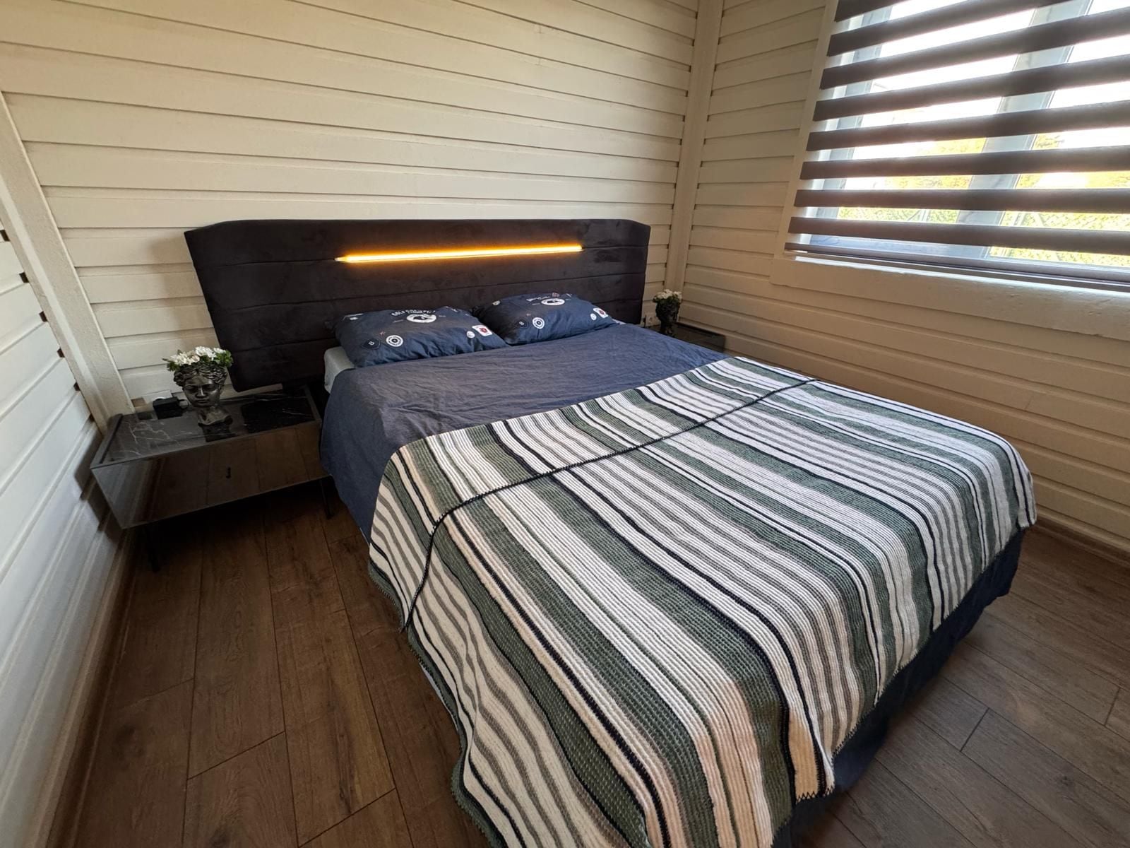 A bedroom with a bed, nightstands, a bedside table, a window with blinds, and a headboard with a light strip.