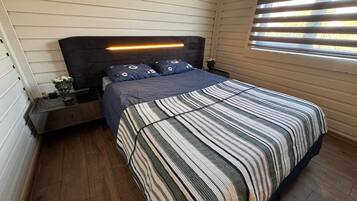 A bedroom with a bed, nightstands, a bedside table, a window with blinds, and a headboard with a light strip.