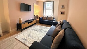 A modern living room with a sectional sofa, a flat-screen TV, a wooden console table, and a large window providing natural light. - Beautifully Presented Cosy Home in Kirkcudbright - Pass the Keys (Kirkcudbright)