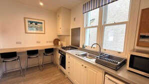 A compact kitchen with a gas stove, sink, and a dishwasher. There are two bar stools and a framed picture on the wall. - Beautifully Presented Cosy Home in Kirkcudbright - Pass the Keys (Kirkcudbright)
