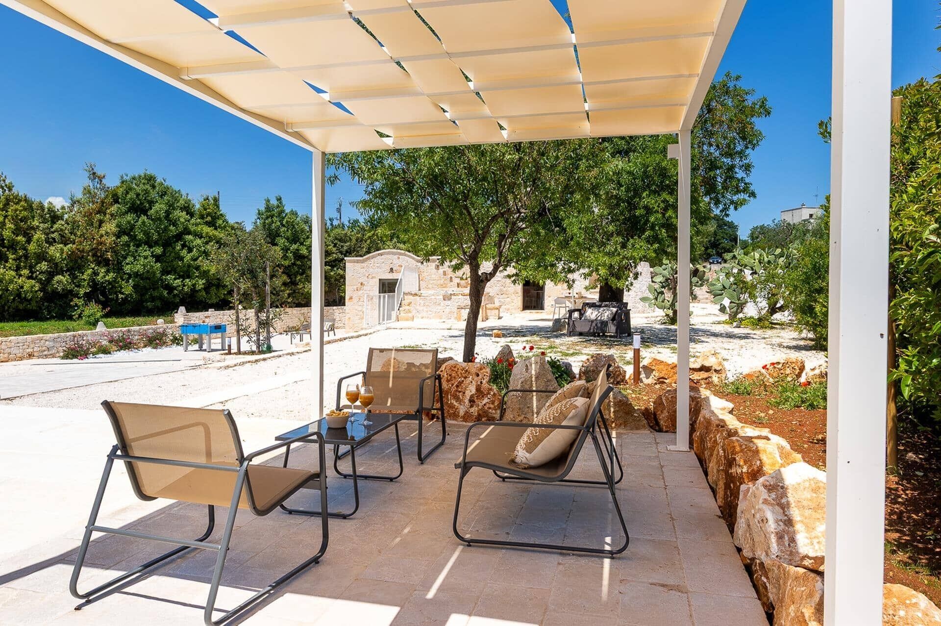 An outdoor seating area with a table and chairs, a pergola overhead, and a stone wall in the background.