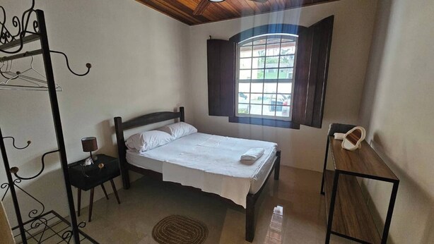 Una cama individual con cabecera de madera, una mesita de noche con lámpara, una ventana con cortinas y un perchero.