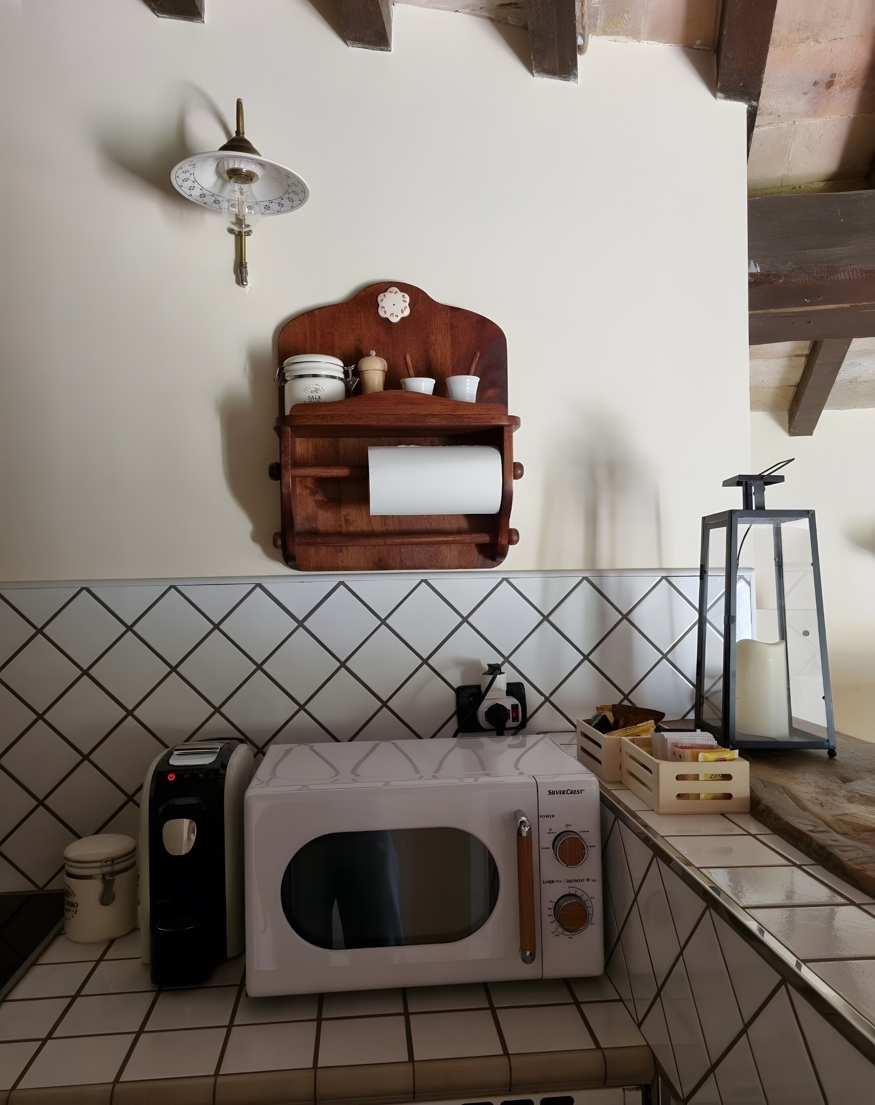 A kitchen with a microwave, a toaster, a wooden shelf with kitchen items, and a wall-mounted light fixture.