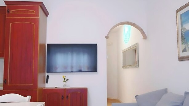 Una sala de estar con un sofá, un televisor fijado a la pared y piso de madera.