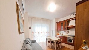Un espacio habitable compacto con un sofá gris, una mesa de comedor con sillas, una zona de cocina con armarios de madera y una lámpara de techo blanca.
