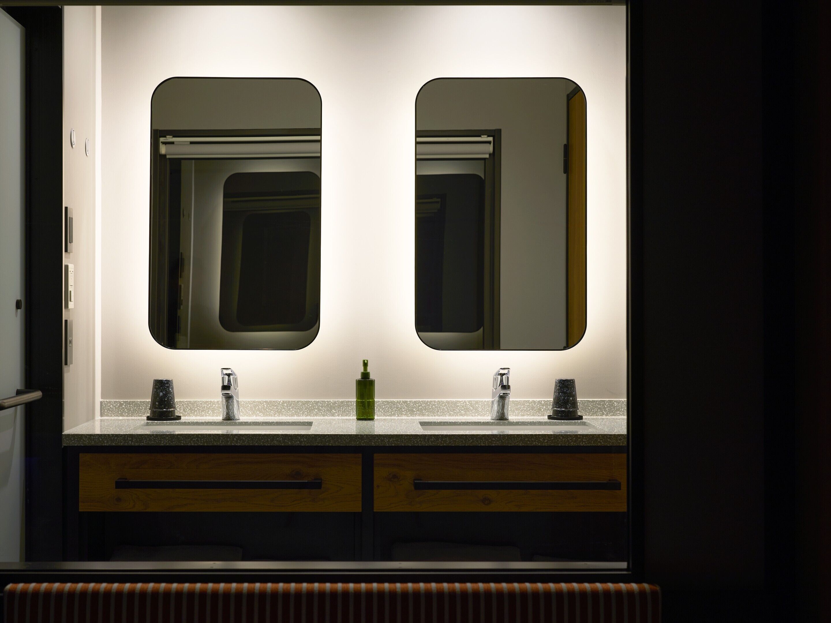 A bathroom with two sinks, each with a mirror above. There are two towel racks, a soap dispenser, and a faucet. The countertop is speckled, and there are wooden drawers below the sinks.