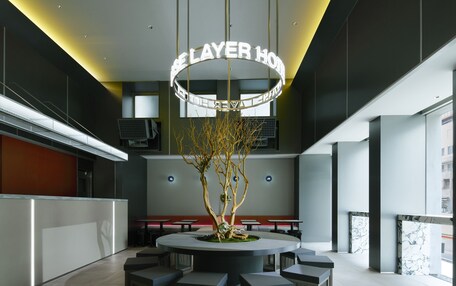 A modern hotel lobby with a central seating area, a decorative tree, and a neon sign reading 'LAYER HOTEL'.