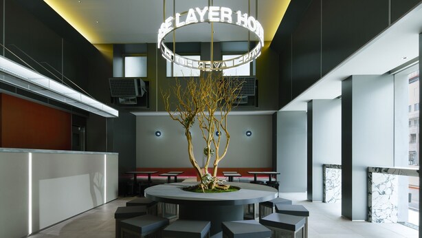 A modern hotel lobby with a central seating area, a decorative tree, and a neon sign reading 'LAYER HOTEL'.