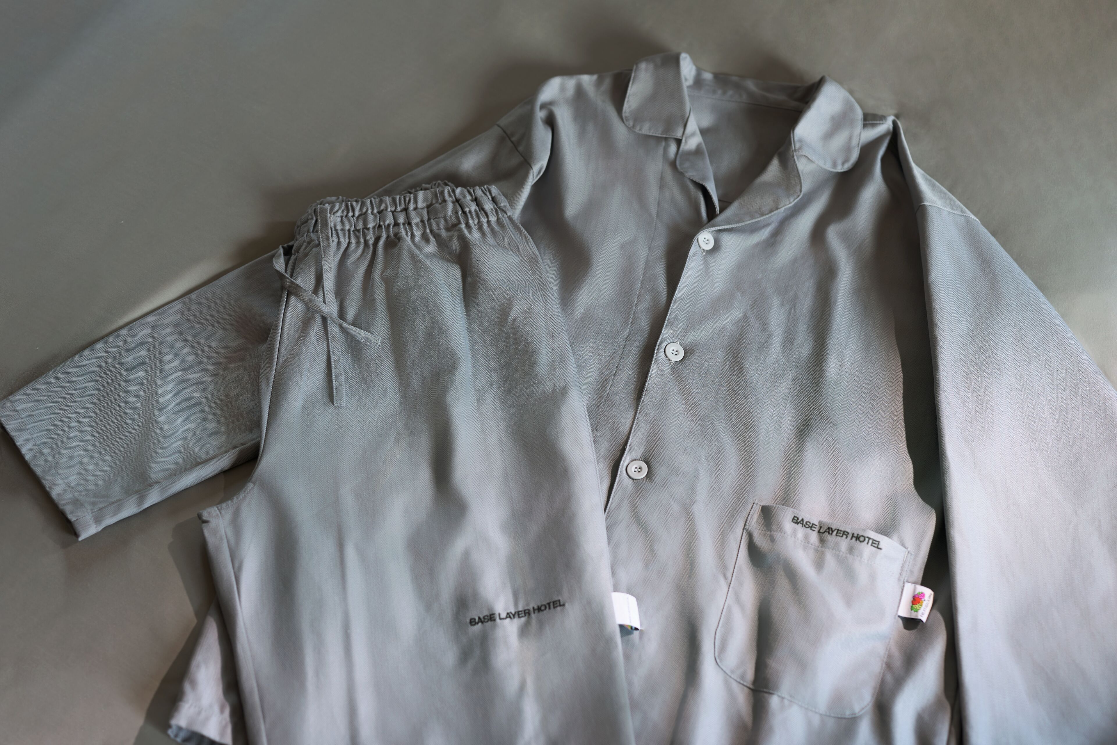A light grey shirt with a collar, short sleeves, and two pockets, with a tag reading 'BASE LAYER HOTEL.'