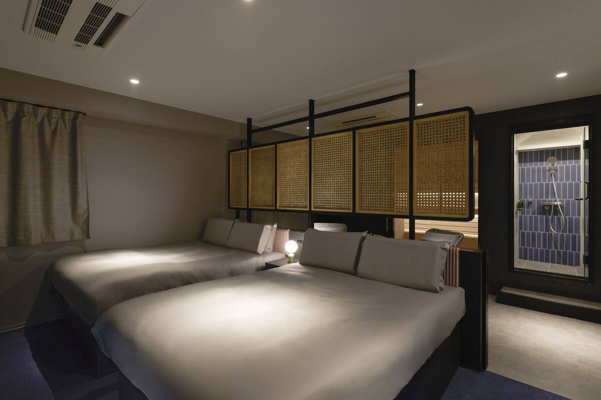 A hotel room with two beds, a window with a view, a bathroom with a shower, and a ceiling vent.